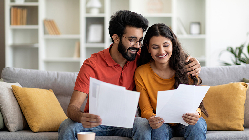 Happy young indian couple checking documents at home, smiling eastern spouses reading insurance agreement or property certificate, man and woman sitting on couch in cozy living room, free space